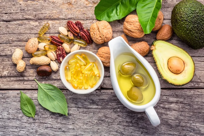 Holztisch mit einer Auswahl an gesunden Fetten, darunter Nüsse (Walnüsse, Mandeln, Pekannüsse, Pistazien, Erdnüsse), Olivenöl in einer weißen Kanne mit Oliven, Avocadohälfte, grüne Blätter und gelbe Omega-3-Kapseln.