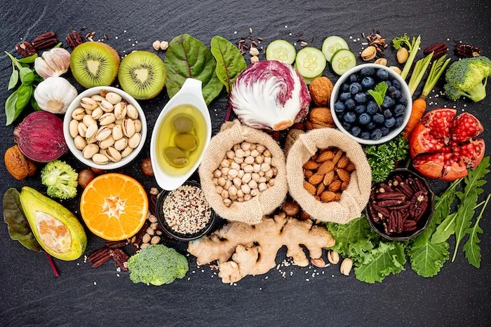 Vielfältige Auswahl an pflanzlichen Lebensmitteln auf schwarzem Untergrund, darunter Kiwi, Knoblauch, Pistazien, Olivenöl, Radicchio, Blaubeeren, Granatapfel, Brokkoli, Avocado, Orange, Quinoa, Kichererbsen, Mandeln, Pekannüsse, Ingwer, Gurkenscheiben, Karotten, Rote Bete und frische Kräuter.